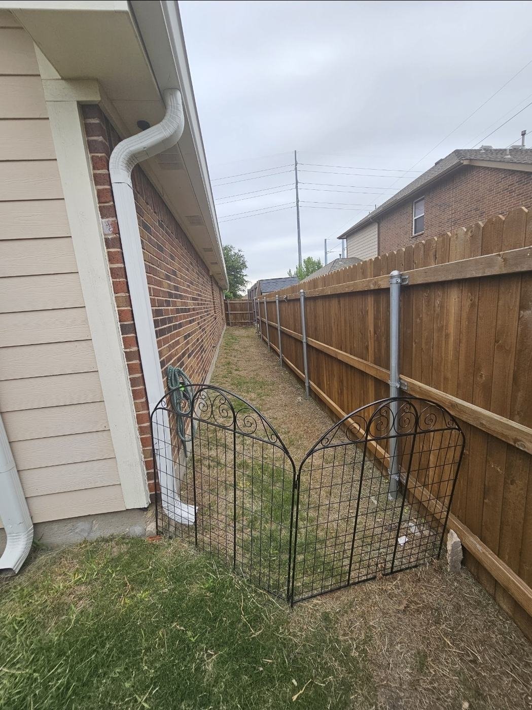 Narrow bare-dirt side yard between a house wall and fence before drainage work