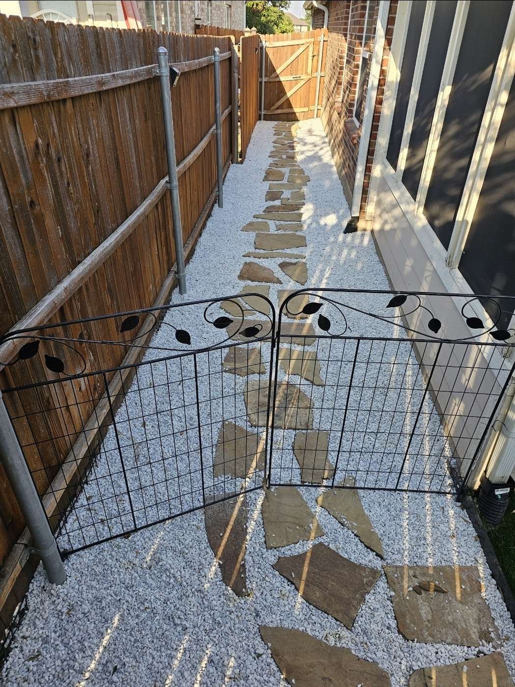 Gravel side yard with a flagstone path and garden gate in afternoon sun, over a French drain